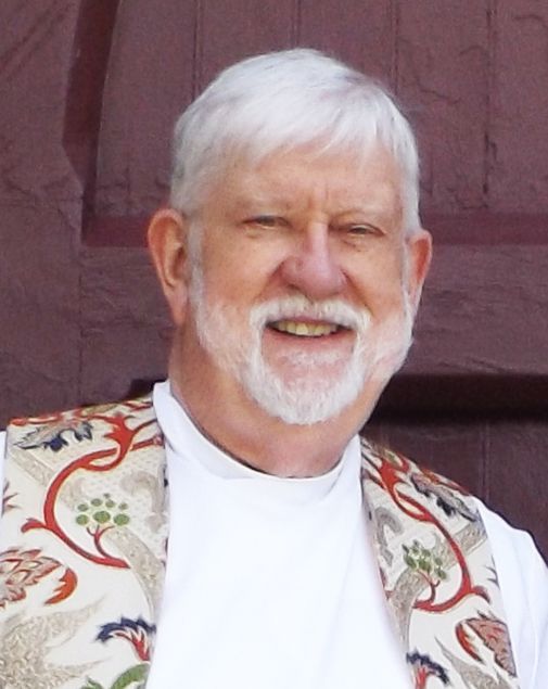 Smiling white-haired Pastor Chuck in a white robe & patterned vestments stands against a red wooden door