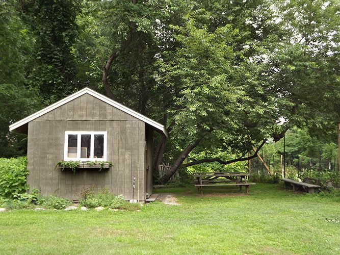 A small gray shed sits in the lush green rear churchyard, w/trees & a picnic table nearby