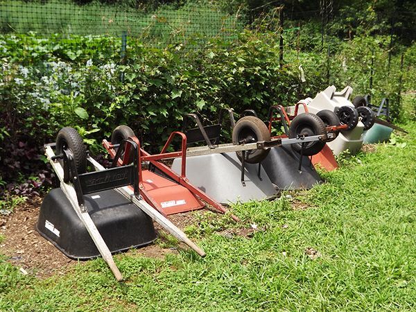 A row of overturned wheelbarrows is lined up in the grass, beside a garden delimited by a netted fence