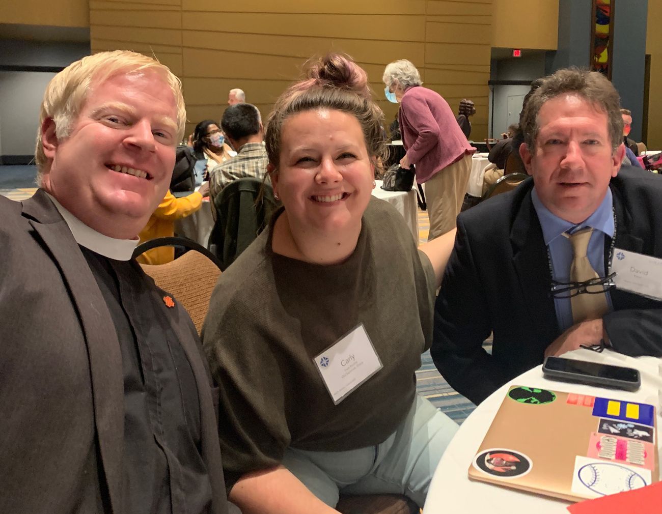 The Pastor sits smiling at a table for the 2022 Convention w/our 2 convention delegates.