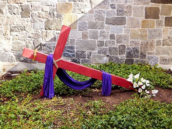 A red wooden cross draped with a purple cloth rests diagonally on its side in the grass beside a stone wall and white flowers