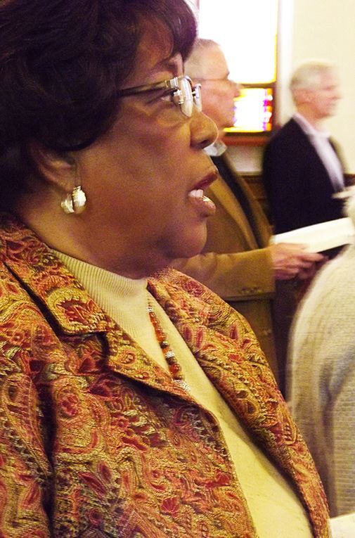 A black woman in earrings & a patterned jacket sits in a pew during worship