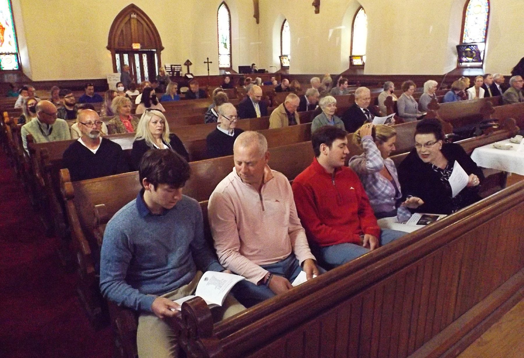 Parishioners seated in wooden pews at church, waiting for worship service to begin, some reading from books.
