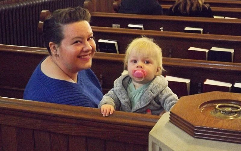 A smiling woman in a purple sweater holds a child with a pink pacifier in a wooden church pew