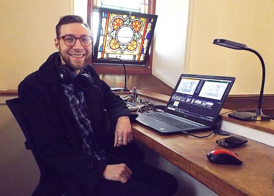 A smiling male Tech-Team volunteer sits at a desk with a laptop, desk lamp, and stained-glass window in the background
