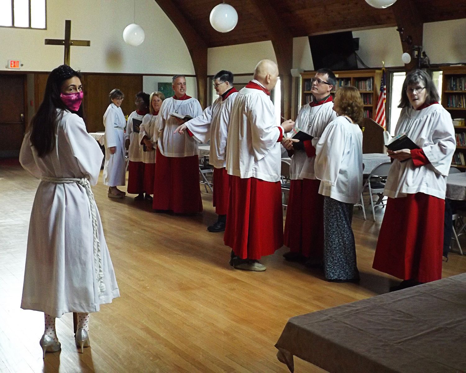 Choir members in white robes & red skirts stand in the Parish Hall with an acolyte holding a cross before worship service