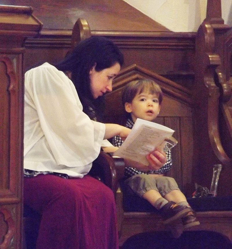 A robed woman shows a worship bulletin to a child seated beside her on a wooden bench in church.
