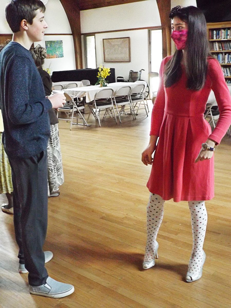 A boy in a dark track-suit talks to a girl in a red dress & white polka-dot tights, while standing in the church Parish Hall.
