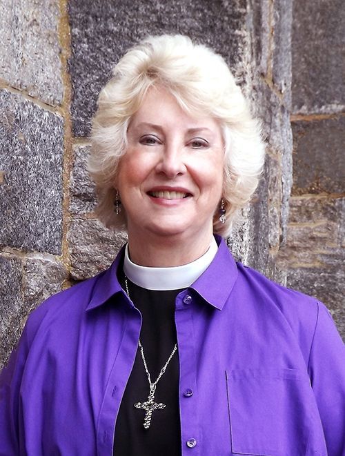 Smiling Pastor Ellendale in a purple shirt and clerical collar stands n front of the church's stone wall