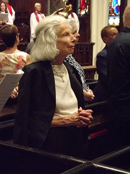 A regal elderly woman with coiffed white hair stands with folded hands in a church pew during a service