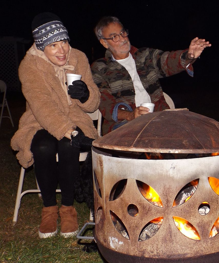 Two bundled people sit by a fire pit at night, one gesturing while flames glow through the sides of the metal fire ring.