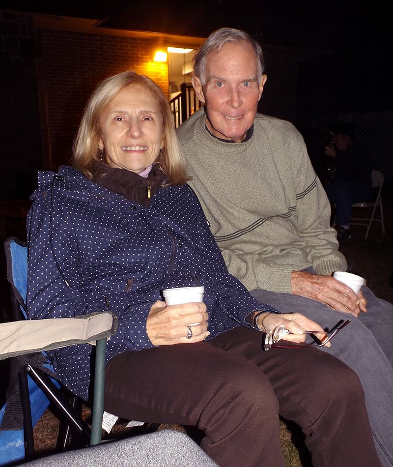 A couple sits together outdoors at night, smiling and holding cups in folding chairs.