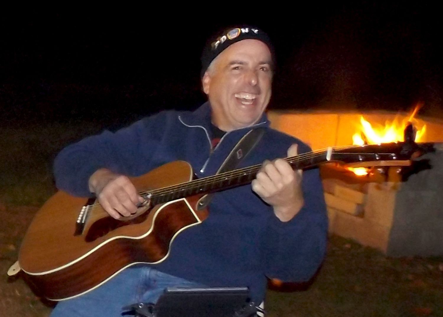 Smiling male parishioner playing acoustic guitar outdoors at night beside a fire pit