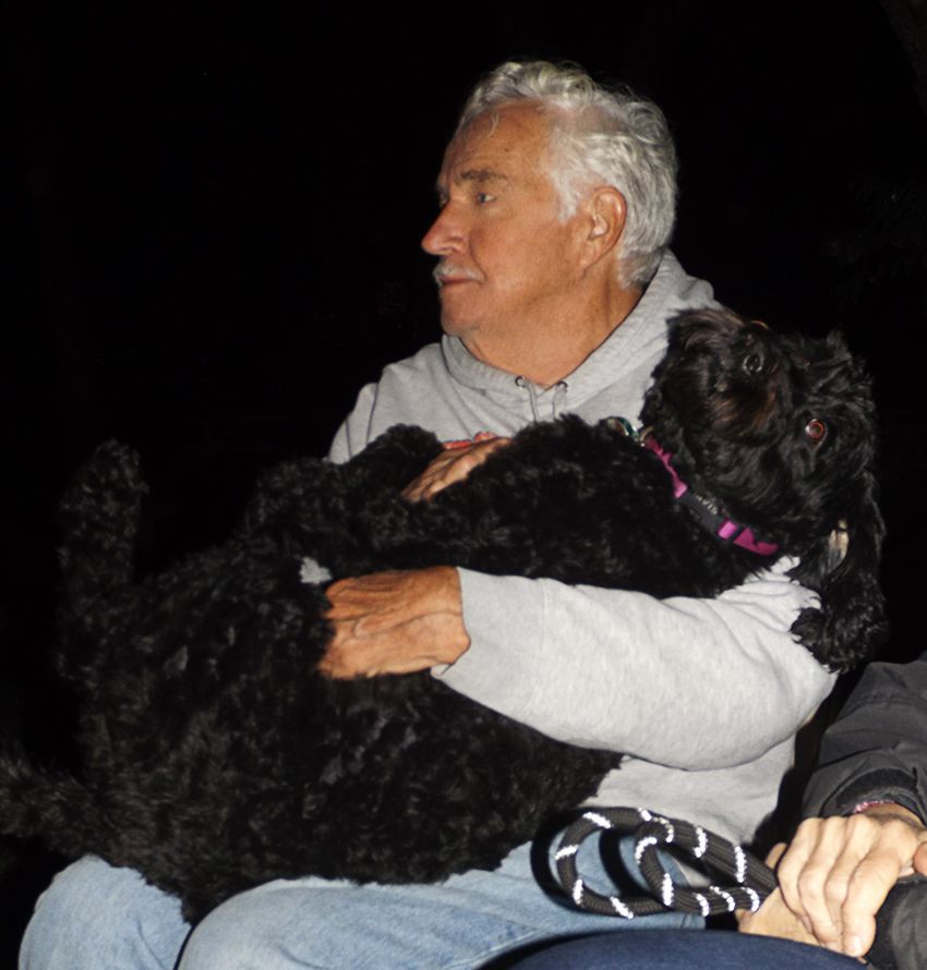 An older man in a gray hoodie holds a large, black, pink-collared dog on his lap.