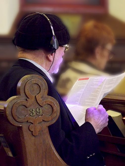 An elderly man in a dark hat, suit and headphones intently reads a bulletin while sitting in a wooden church pew