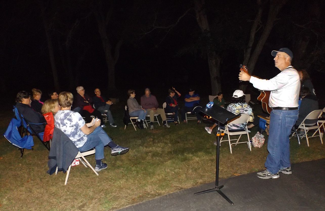 Evening firepit group outdoors with a parishioner-musician playing guitar and singing to seated attendees around a campfire