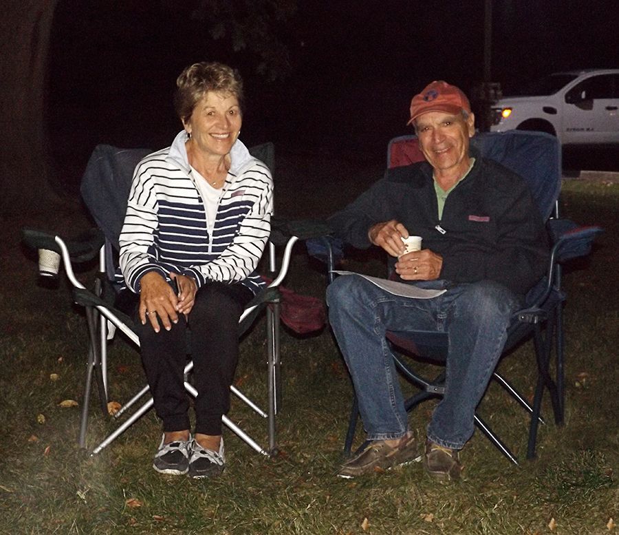 A couple sits smiling side by side in camping chairs outside in the churchyard at night, w/a vehicle in the background.