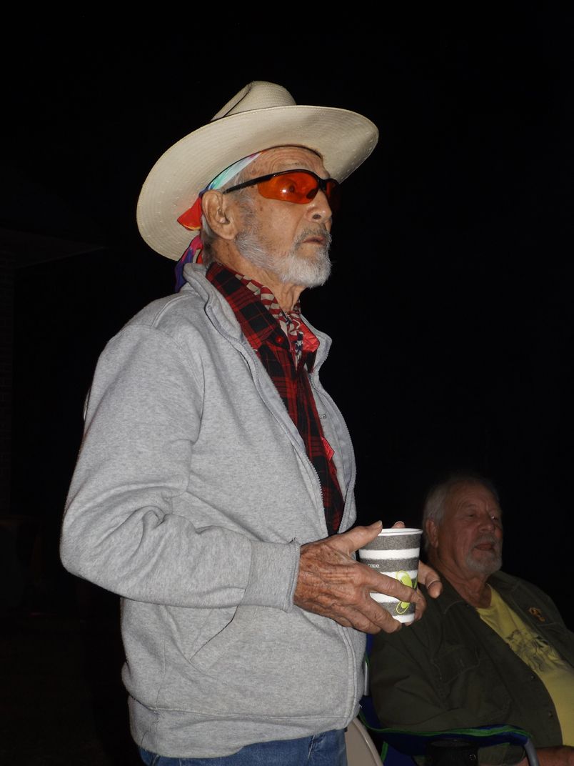 Man in a cowboy hat and red glasses holds a cup outdoors at a nighttime firepit event.