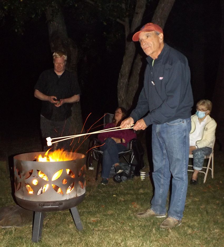 Male parishioner roasting marshmallows over a fire pit outdoors at night while others sit nearby