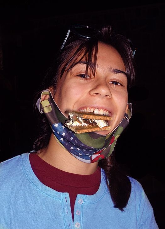 A smiling girl in a blue shirt balances a s'more treat in her mouth, against a dark nighttime background.
