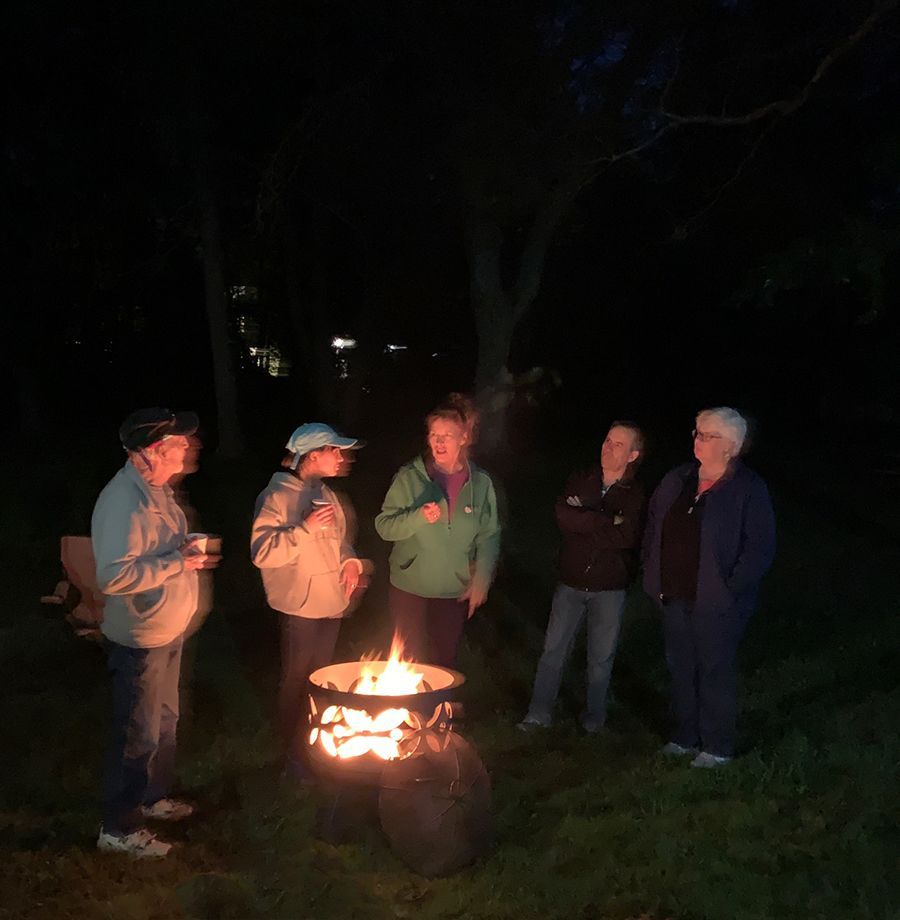 5 people stand around a small campfire chatting at night in the dark churchyard.