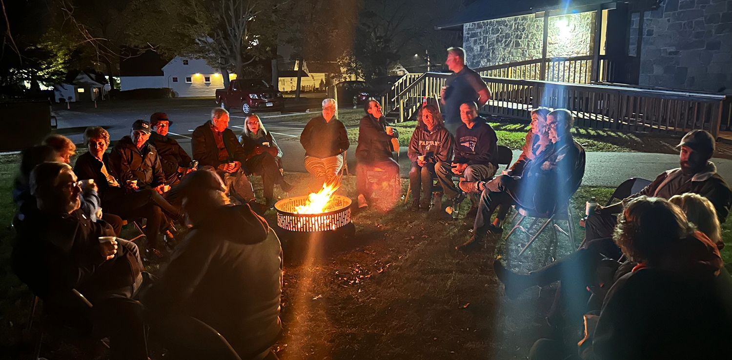 People cluster together in a circle of chairs around a fire pit at night, with a lit building nearby