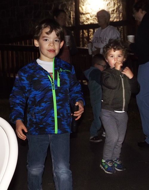 2 little boys stand outdoors in the semi-dark, one wearing a blue camo jacket & the other a gray hoodie sucking his thumb.