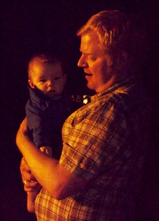 The Pastor holds a baby outdoors at night, near the amber glow of the fire