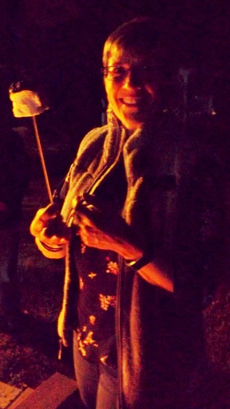 A woman stands smiling while holding a lit marshmallow on a stick near a firepit at night.