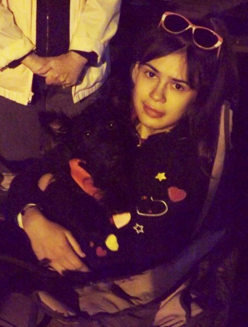 A young woman wearing a black sweater w/colorful heart patterns sits outdoors holding a Black Scottish Terrier dog in her lap