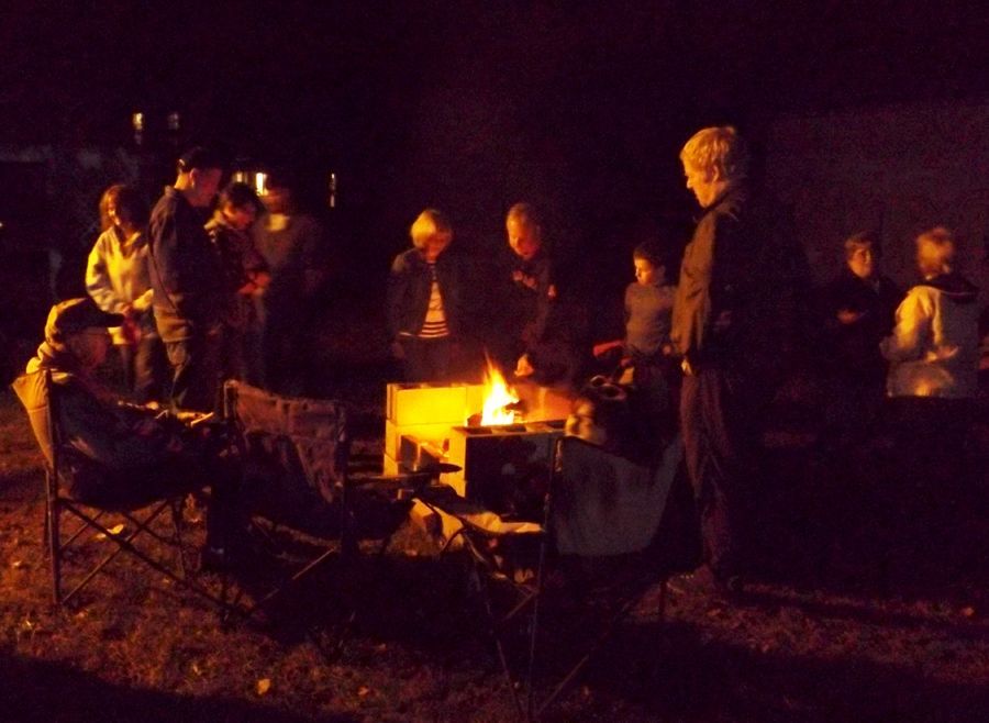 People gather around a campfire at night, w/chairs & a glowing fire in the dark.