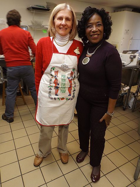 Two women stand together in the Church kitchen, one wearing a red sweater & apron, the other in dark purple w/a necklace