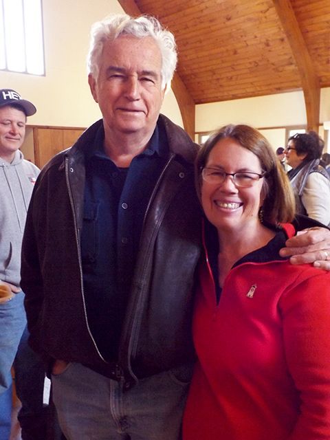 A man  in a black jacket & woman in a red jacket stand smiling together in the Parish Hall