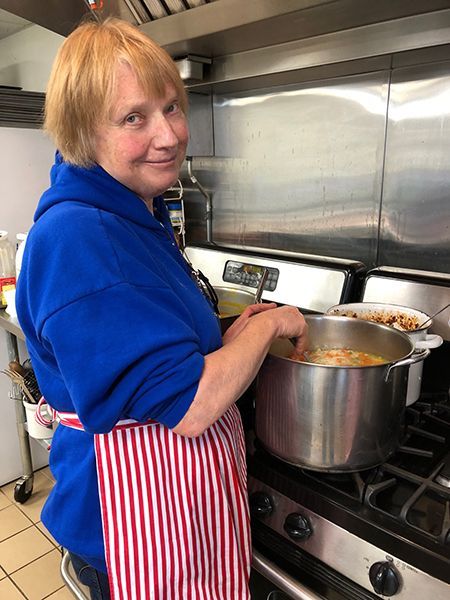 A woman in a blue hoodie & red-striped apron looks over her shoulder while stirring a large pot in a commercial kitchen.