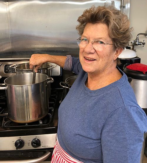 A short-haired woman in a greyish blue shirt & a red & white striped apron stirs a pot on a stove in the Church kitchen