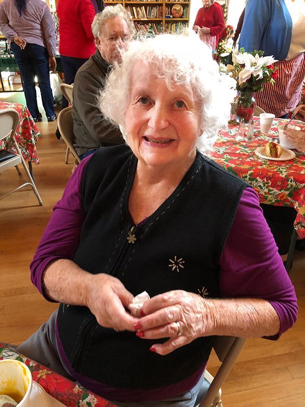 Elderly woman in a black vest and purple top sits at a festive table with flowers and decorations.