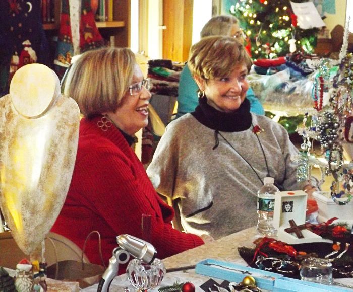Two women sit smiling in red & gray sweaters at a table with a festive holiday craft display during the church Holiday Fair.