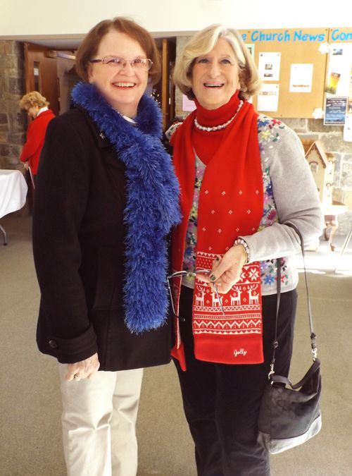 In the church entry foyer 2 women smile, wearing festive scarves, while standing by a bulletin board.