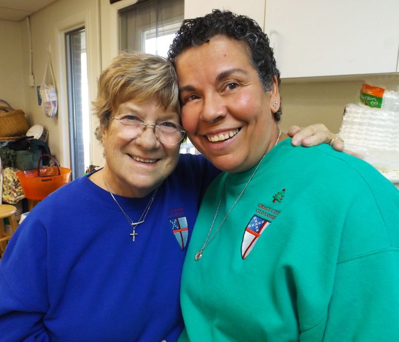 2 smiling women in blue & green sweatshirts w/Church emblems stand close together in the Church kitchen.