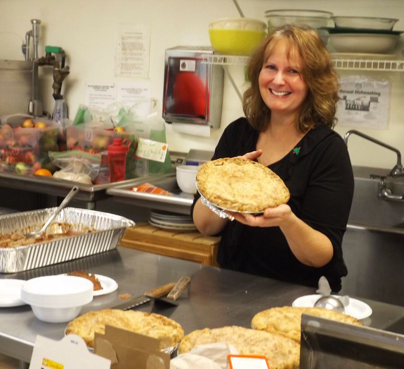 Woman in church kitchen smiling while holding a freshly baked pie over a counter, surrounded by more pies.