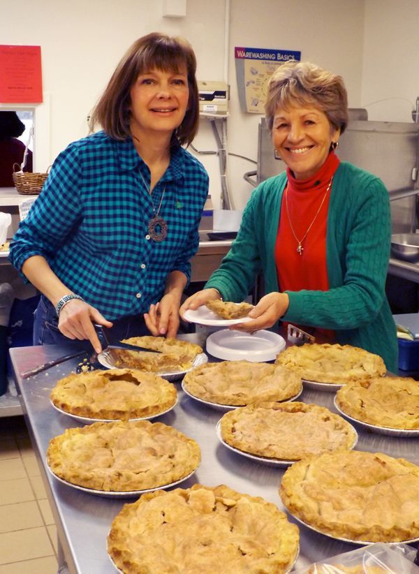 Two smiling women serving pies from a table in the church kitchen area.
