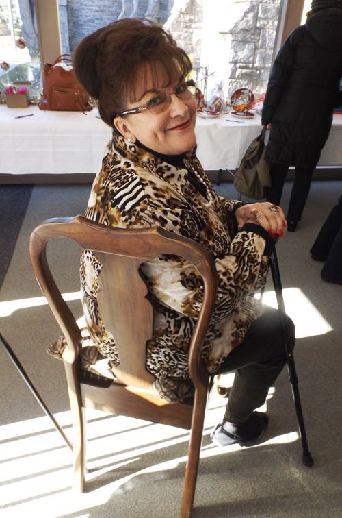 A dimpled, seated woman with a cane looks over her shoulder smiling, in a leopard-print blouse, in the sunlit church foyer.