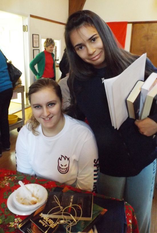 Two smiling young adult female friends: one seated at a table with a bowl of soup, and one standing above her, holding books.