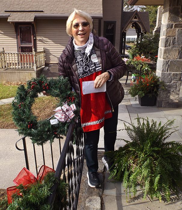 A woman in sunglasses stands in a dark jacket & red apron, holding a decorated wreath on the church's stone patio
