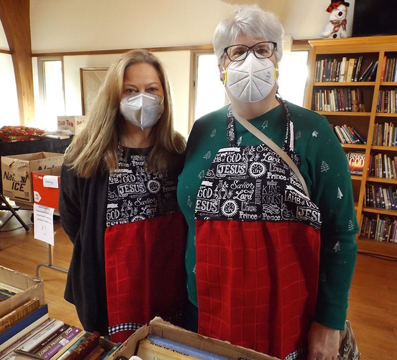 Two masked women in black-&-red aprons stand in the Parish Hall among tables & bookshelves