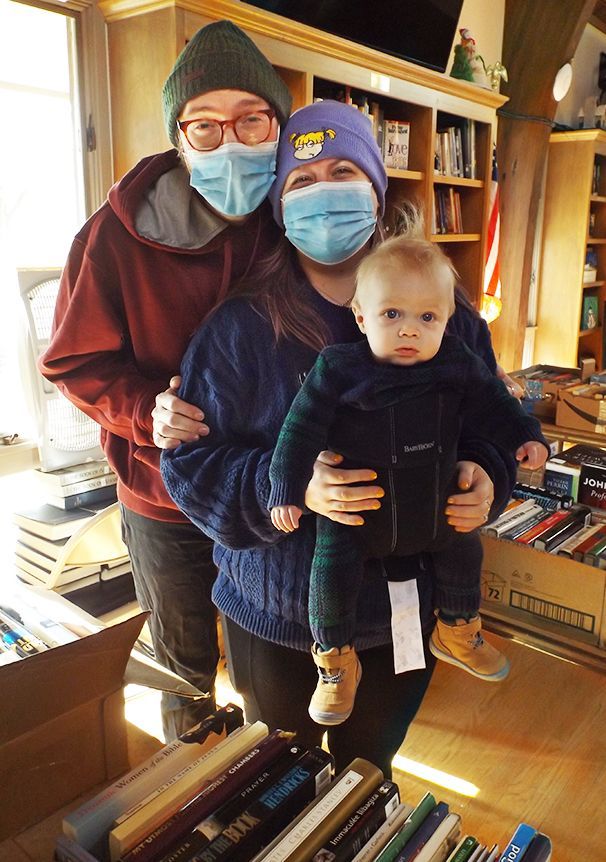 Two masked parents wearing hats and coats, hold their baby son in the cozy Parish Hall, amidst books and warm sunlight