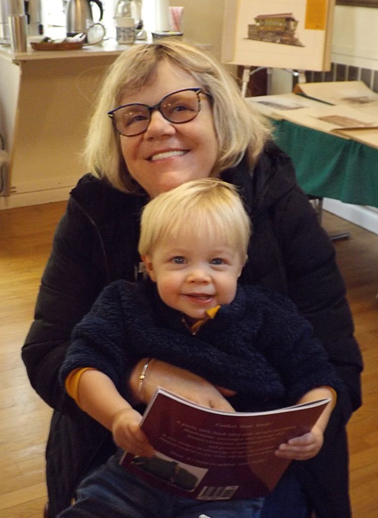 A smiling blond woman wearing glasses embraces a blond toddler boy who is holding a book in the warmly-lit Parish Hall