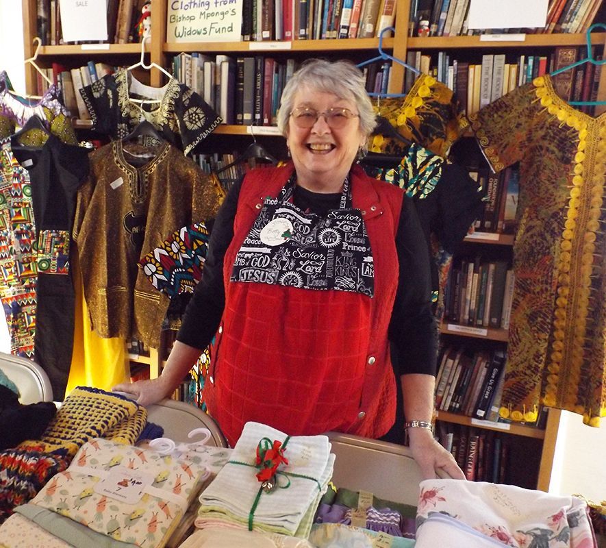 Smiling woman vendor behind a table of folded fabric crafts, with colorful brocaded garments hanging behind her