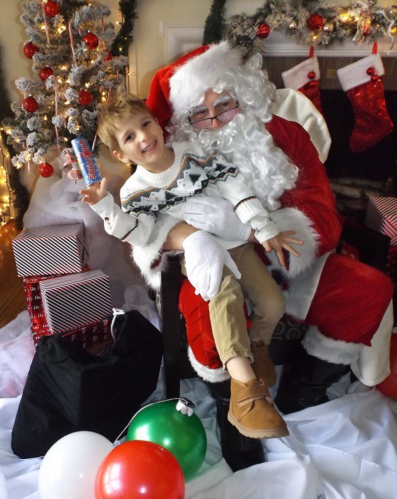 Santa Claus holds a smiling, wiggly young boy on his lap, in a festively decorated room