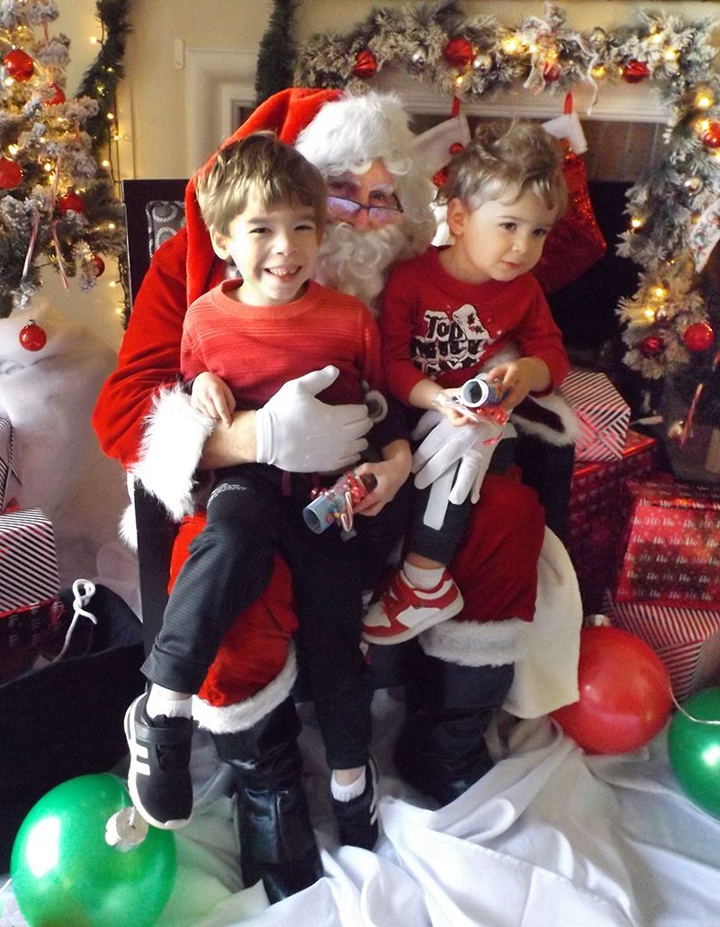 Two young boys in red shirts sit on Santa’s lap in a decorated holiday room